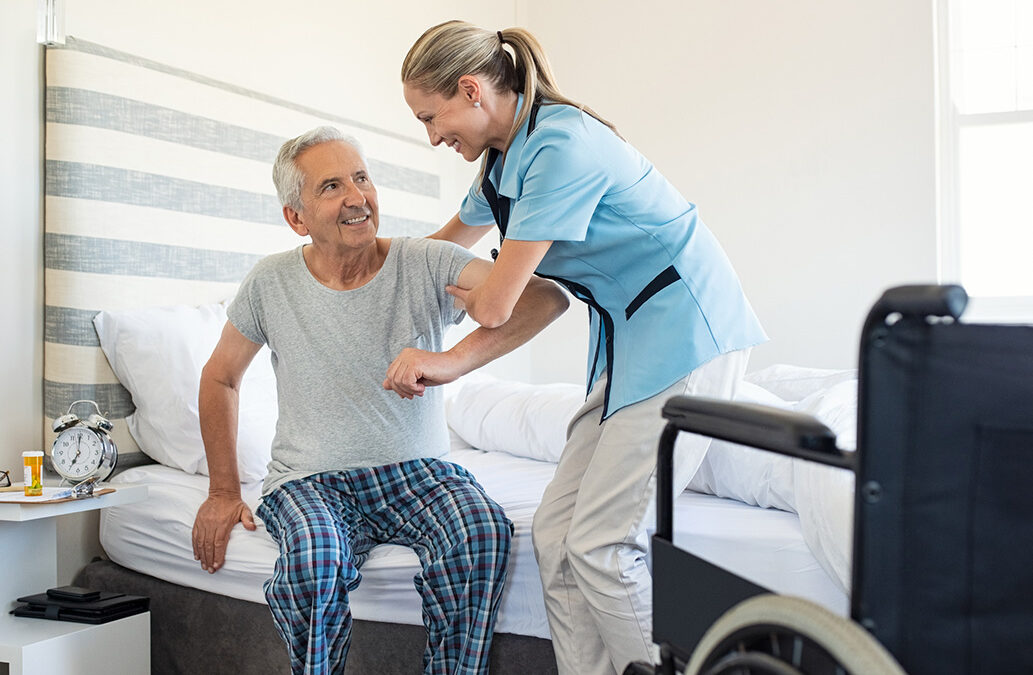 Smiling nurse assisting senior man to get up from bed. Caring nurse supporting patient while getting up from bed and move towards wheelchair at home. Helping elderly disabled man standing up in his bedroom.