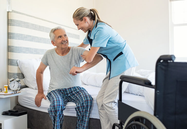 Smiling nurse assisting senior man to get up from bed. Caring nurse supporting patient while getting up from bed and move towards wheelchair at home. Helping elderly disabled man standing up in his bedroom.