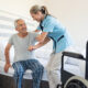 Smiling nurse assisting senior man to get up from bed. Caring nurse supporting patient while getting up from bed and move towards wheelchair at home. Helping elderly disabled man standing up in his bedroom.