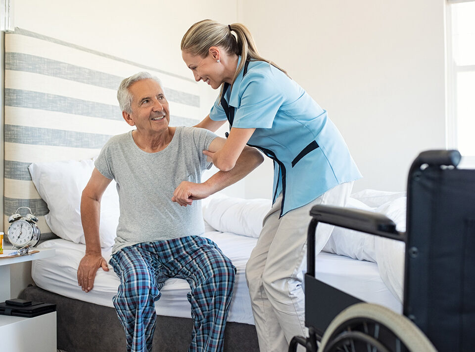 Smiling nurse assisting senior man to get up from bed. Caring nurse supporting patient while getting up from bed and move towards wheelchair at home. Helping elderly disabled man standing up in his bedroom.